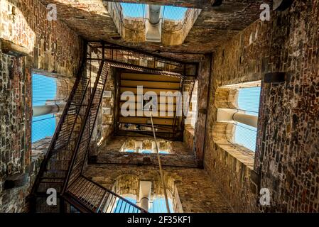 Steel stairway around walls of square tower with windows. View up from down side. Natural light inside building. Stock Photo