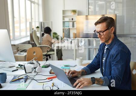 Side view portrait of tattooed IT developer designing VR software while using computer and working in office, copy space Stock Photo