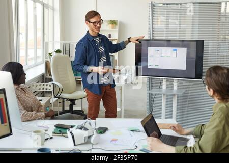 Meeting of IT development team in office with focus on tattooed man standing by digital screen and giving presentation, copy space Stock Photo