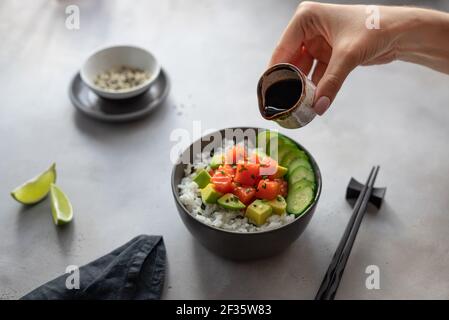 woman's hands holding a saucepan with soy sauce to sprinkle a Hawaiian poke bowl with salmon, rice, avocado and cucumber. traditional asian food Stock Photo