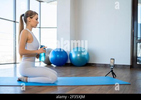 Athletic girl with bottle in hand sitting on mat in front of the camera phone and recording her fitness blog. Yoga instructor doing online classes Stock Photo