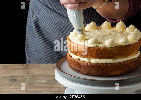 A baker adds butter cream to the top of a freshly home baked cake. The baker, a female is skillfully adding the butter cream using a piping bag Stock Photo