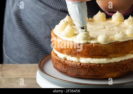 A baker adds butter cream to the top of a freshly home baked cake. The baker, a female is skilfully adding the butter cream using a piping bag Stock Photo