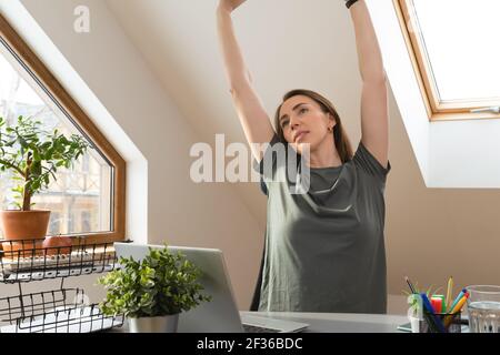 Woman stretching her hands up at office desk in a break from work at home office. Exercise and healthy living concept. Stock Photo
