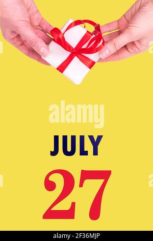 July 27th. Festive Vertical Calendar With Hands Holding White Gift Box With Red Ribbon And Calendar Date 27 July On Illuminating Yellow Background.Sum Stock Photo