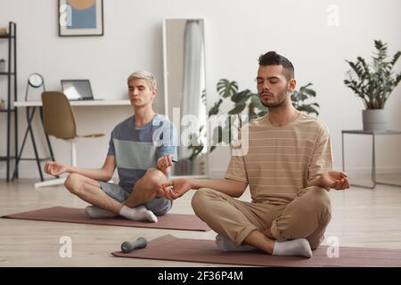 Full length portrait of two young men meditating at home while enjoying yoga exercises sitting on mats in lotus position Stock Photo