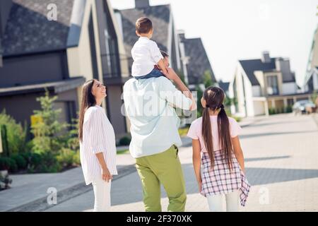 Back rear spine view photo of happy cheerful smiling family walk outdoors dad carry son on shoulders mom and school girl watch Stock Photo