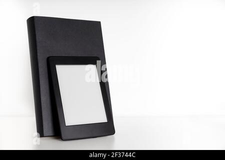 Black book with blank spine and front cover and an electronic reader on a white shelf with white background. Concepts of technology, reading and educa Stock Photo
