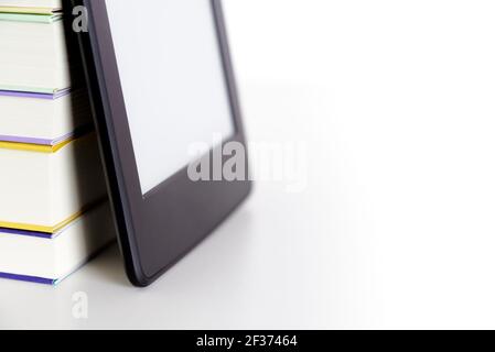 Corner of a stack of books and an electronic reader with a blank screen leaning against them, on a white surface. Concepts of technology, reading and Stock Photo