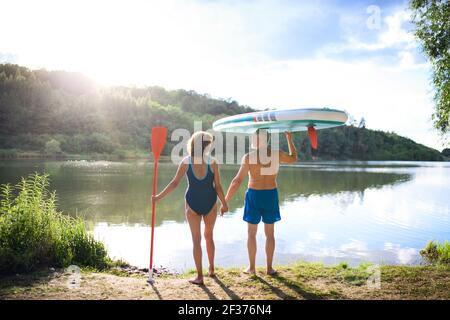 Rear view of senior couple carrying paddleboard by lake in summer. Stock Photo