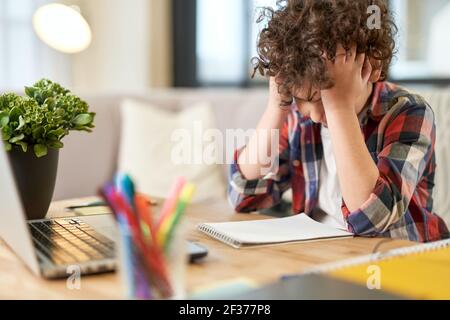 Fatigue. Portrait of tired latin boy holding his head, looking sad while sitting at the desk and doing homework at home Stock Photo