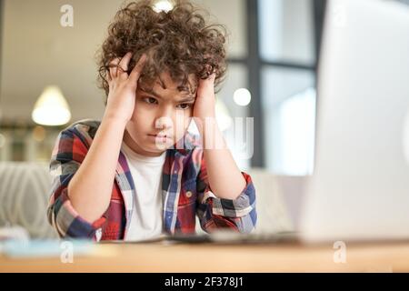Tired kid. Portrait of tired latin boy holding his head, looking sad while studying at home using laptop and sitting at the desk indoors Stock Photo