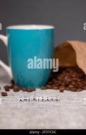 blue cup near coffee beans falling from paper bag and word AMERICANO written with little white cubes in front. Stock Photo