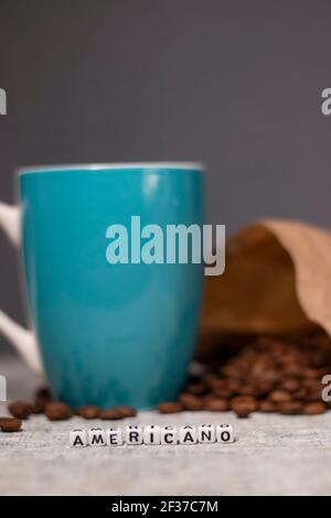 blue cup near coffee beans falling from paper bag and word AMERICANO written with little white cubes in front. Stock Photo
