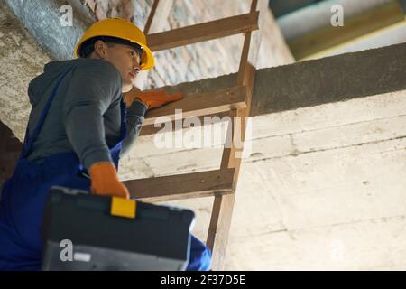 Young male builder in blue overalls and hard hat looking focused, climbing up the ladder, holding toolbox while working at construction site Stock Photo