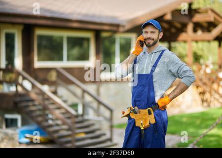 Cheerful young male builder in uniform smiling at camera while talking on the phone, standing outdoors at cottage construction site on a warm sunny Stock Photo