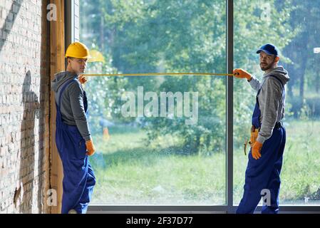 Two positive young male builders wearing blue overalls looking at camera, using measuring tape while working on cottage construction site Stock Photo