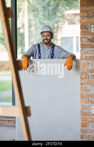 Handsome young male builder in hard hat smiling at camera, holding drywall while working at construction site Stock Photo