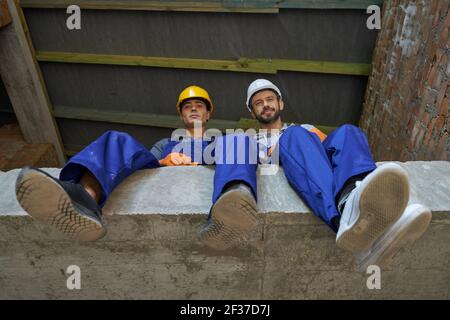 Two attractive young builders in blue overalls and hard hats looking down at camera, sitting on the concrete floor, resting while working on house Stock Photo