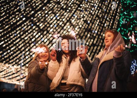 Happy female friends holding sparklers standing by christmas lights Stock Photo