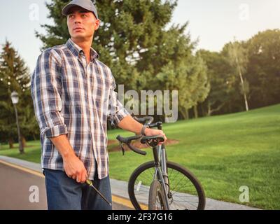 Handsome caucasian man looking away, holding a screwdriver for repairing his bike, standing in the public park Stock Photo