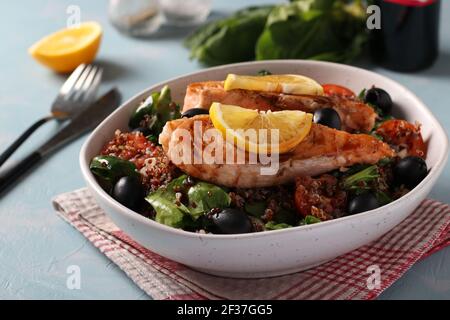 Salad with quinoa, salmon, spinach, black olives, lemon and cherry tomatoes in a plate on light blue background. Stock Photo