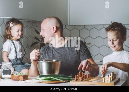 Father whips an omelet with a whisk, playing chess with son and talking to daughter. Man doing household chores. Stock Photo