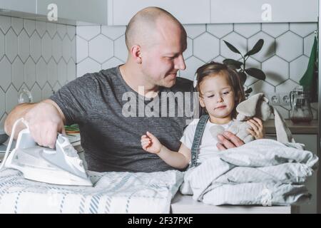 Man ironing bed linen with little daughter on his knees. Father engaged in household chores. Stock Photo