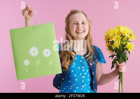 happy stylish girl in dotted blue overall with yellow chrysanthemums flowers and green shopping bag isolated on pink background. Stock Photo