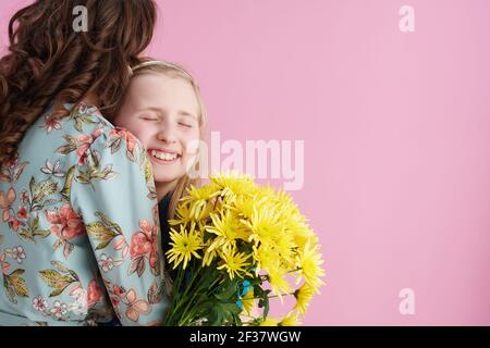 happy elegant mother and child with long wavy hair with yellow chrysanthemums flowers embracing isolated on pink. Stock Photo