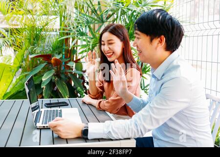 Cheerful young Asian man and woman waving hands sitting in cafe while using laptop for video call with friends and family during covid-19 Stock Photo