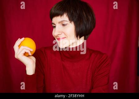 Smiling mature brunette woman looking at orange fruit in hand, close-up face portrait. Generation X,  Stock Photo