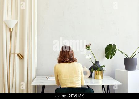Back view of anonymous female sitting at table with flowers in pots and vase in room in minimal style Stock Photo
