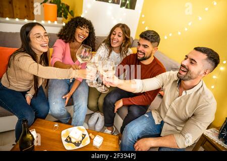 Team of young happy people celebrating at home toasting with champagne wine glasses sitting at the sofa in the living room. Millennial student drinkin Stock Photo