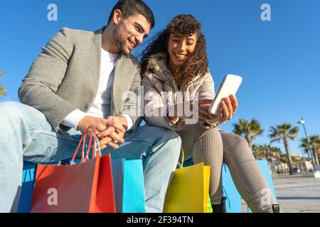 Mixed race young couple of millennial using internet mobile technology for online shopping outdoor. Happy people having fun sitting on city park banch Stock Photo