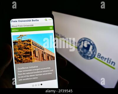 Person holding cellphone with website of German university Freie Universität Berlin (FU) on screen in front of logo. Focus on center phone of display. Stock Photo