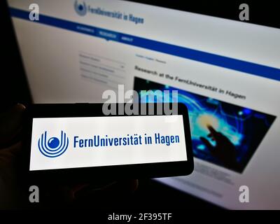 Person holding cellphone with logo of German education institution University of Hagen on screen in front of webpage. Focus on phone display. Stock Photo