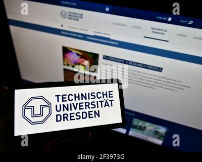 Person holding smartphone with logo of German university Technische Universität Dresden (TUD) on screen in front of web page. Focus on phone display. Stock Photo