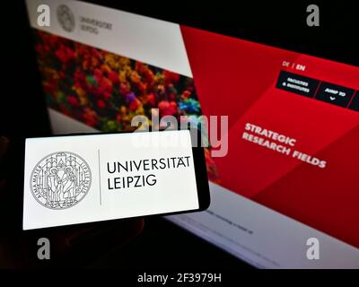 Person holding smartphone with logo of German education institution Leipzig University on screen in front of webpage. Focus on phone display. Stock Photo