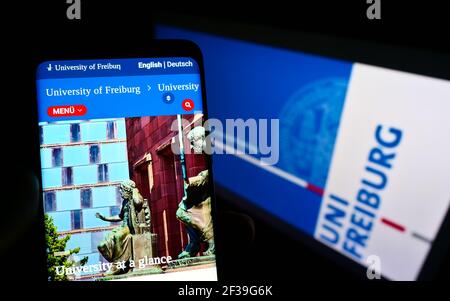 Person holding smartphone with webpage of German University of Freiburg on screen in front of logo. Focus on top-left of phone display. Stock Photo