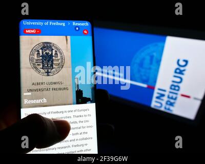 Person holding mobile phone with webpage of German University of Freiburg on screen in front of logo. Focus on center of cellphone display. Stock Photo