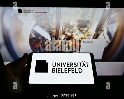 Person holding cellphone with logo of German education institution Universität Bielefeld on screen in front of webpage. Focus on phone display. Stock Photo