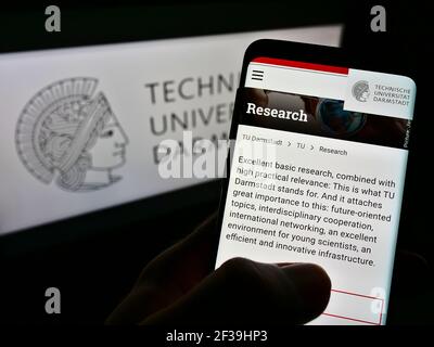 Person holding cellphone with webpage of German Darmstadt University of Technology on screen in front of logo. Focus on center of phone display. Stock Photo