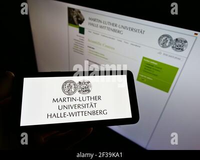 Person holding cellphone with logo of German Martin Luther University of Halle-Wittenberg  on screen in front of webpage. Focus on phone display. Stock Photo