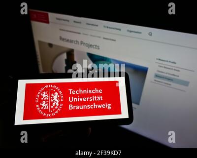 Person holding smartphone with logo of German university Technische Universität Braunschweig on screen in front of webpage. Focus on phone display. Stock Photo