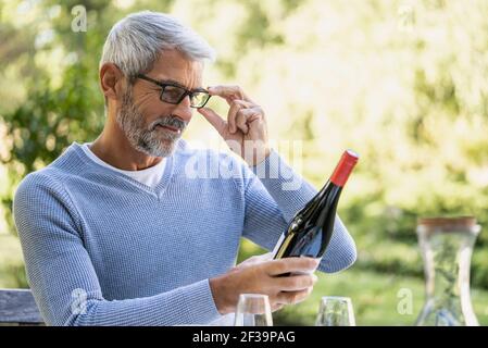 Mature man looking at wine bottle while sitting on chair Stock Photo