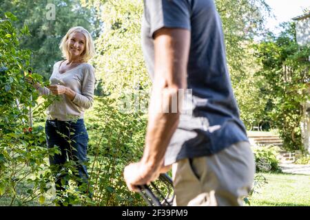 Smiling mature woman with secateurs looking at her husband in backyard Stock Photo
