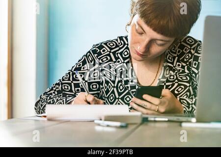 Woman using smartphone while writing in notepad at home Stock Photo