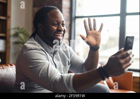 Joyful african american freelancer guy, uses mobile phone, have video call, communicate with colleagues or friends by video conference, waving hand, smiling. Handsome black man working from home Stock Photo