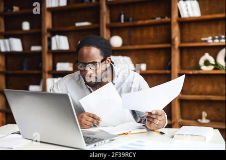 Young concentrated manager sitting in cabinet surrounded with papers documents, accountant analyst having careful look at the laptop screen, doing online trade market tech research, preparing report Stock Photo
